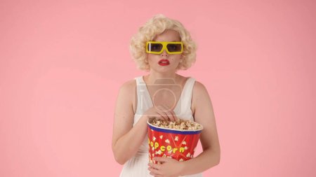 Téléchargez les photos : Femme portant des lunettes 3D et avec un grand seau de pop-corn, profiter de regarder un film. Femme comme Marilyn Monroe en studio sur fond rose - en image libre de droit