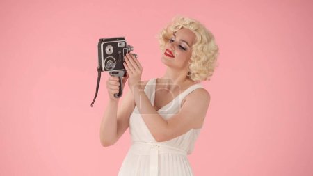 Téléchargez les photos : Femme prenant selfies en utilisant une vieille caméra vidéo vintage. Femme en look Marilyn Monroe pose pour caméra sur fond rose en studio - en image libre de droit