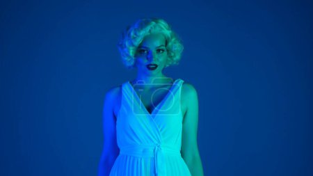 Téléchargez les photos : Portrait d'une femme charmante à l'image de Marilyn Monroe en gros plan. Femme avec maquillage lumineux en robe blanche et perruque en studio en néon rose et vert - en image libre de droit