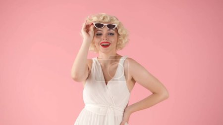 Téléchargez les photos : Femme levant les lunettes de soleil en forme d'oeil de chat vers le haut, souriant. Femme en perruque blanche, robe blanche et rouge à lèvres sur ses lèvres dépeint Marilyn Monroe en studio sur fond rose. Personnes, mode de vie émotionnel - en image libre de droit