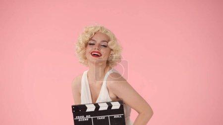 Foto de Mujer en la imagen de Marilyn Monroe sosteniendo una tabla de aplausos. Mujer brillante con lápiz labial rojo en los labios, con una peluca y un vestido blanco en un estudio sobre un fondo rosa - Imagen libre de derechos