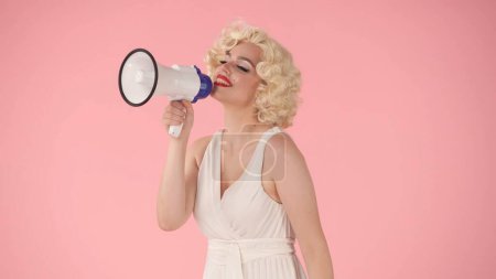 Foto de Mujer en el personaje de Marilyn Monroe hablando en megáfono. Mujer con maquillaje colorido, peluca y vestido blanco en estudio sobre fondo rosa. Venta, Viernes Negro. Copiar espacio - Imagen libre de derechos