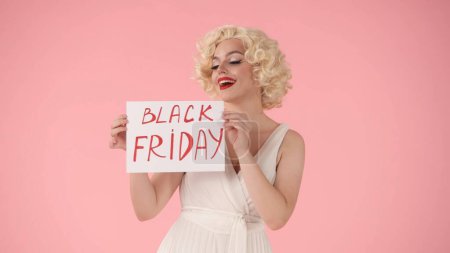 Téléchargez les photos : Femme tenant une affiche avec l'inscription Black Friday. Femme ressemblant à Marilyn Monroe en studio sur fond rose. Vente, vendredi noir, rabais - en image libre de droit