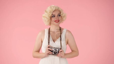 Photo for A woman holds up an old vintage camera. Woman in the image of Marilyn Monroe in studio on pink background close up - Royalty Free Image