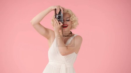 Photo for Woman taking pictures on old camera. Woman looking like Marilyn Monroe in studio on pink background close up - Royalty Free Image
