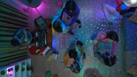 Téléchargez les photos : Groupe d'adolescents danse lente dans une salle de fête décorée avec une balle disco. Un homme mélange de la musique sur une console DJ. Des néons. Vue de dessus. Lycée et collège, publicité ou contenu créatif. - en image libre de droit