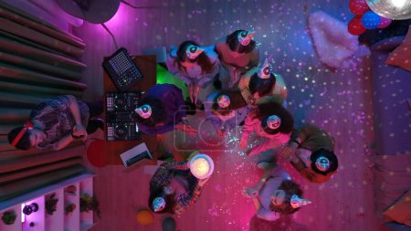 Téléchargez les photos : Groupe d'adolescents célébrant l'anniversaire des filles dans une salle de fête décorée avec un bal disco. Un homme mélange de la musique sur une console DJ. Néon lumières colorées. Vue de dessus. Publicité ou contenu créatif. - en image libre de droit