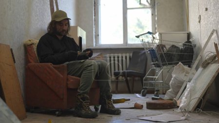 Photo for Homeless poor man sitting in a room of an abandoned building filled with his meager belongings. He is holding a few money bills, notes in his hands. Homelessness and poverty, unemployment, crisis. - Royalty Free Image