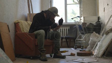 Téléchargez les photos : Pauvre sans-abri assis dans une pièce d'un immeuble abandonné remplie de ses maigres biens. Il s'injecte une substance illégale, de la drogue dans la veine. Sans-abri et pauvreté, chômage, crise. - en image libre de droit