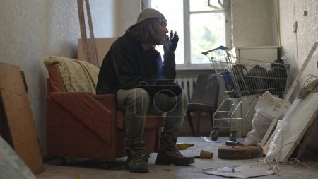Téléchargez les photos : Pauvre sans-abri assis dans une pièce d'un immeuble abandonné remplie de ses maigres biens. Il fume une cigarette ou un joint de cannabis, contondant. Sans-abri et pauvreté, chômage. - en image libre de droit