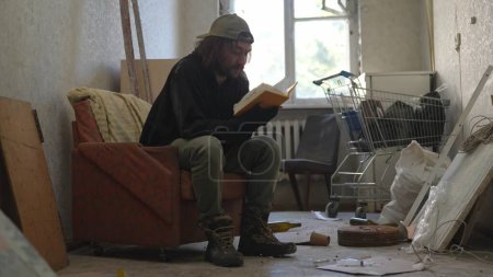 Téléchargez les photos : Pauvre sans-abri assis dans une pièce d'un immeuble abandonné remplie de ses maigres biens. Il lit un livre, feuillette les pages. Sans-abri et pauvreté, chômage, crise. - en image libre de droit