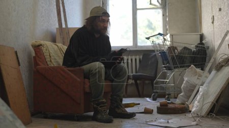 Téléchargez les photos : Pauvre sans-abri assis dans une pièce d'un immeuble abandonné remplie de ses maigres biens. Il écoute la musique à travers les écouteurs. Sans-abri et pauvreté, chômage, crise. - en image libre de droit