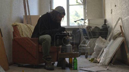 Téléchargez les photos : Pauvre sans-abri assis dans une pièce d'un immeuble abandonné remplie de ses maigres biens. Il chauffe sa nourriture sur un brûleur à gaz. Sans-abri et pauvreté, chômage, crise. - en image libre de droit