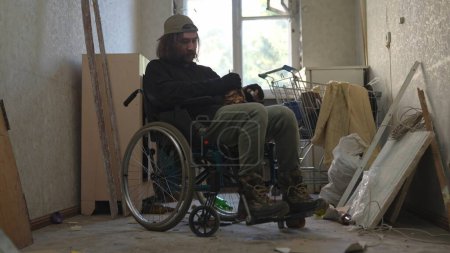 Téléchargez les photos : Un pauvre sans-abri assis dans un fauteuil roulant dans une pièce d'un immeuble abandonné remplie de ses maigres biens. Il tient une bouteille d'alcool. Sans-abri et pauvreté, chômage, crise. - en image libre de droit