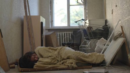Foto de Desamparado pobre hombre durmiendo en un pedazo de cartón, cubierto con una manta en una habitación de un edificio abandonado lleno de sus escasas pertenencias. Desamparo y pobreza, desempleo, crisis. - Imagen libre de derechos