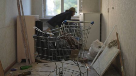 Téléchargez les photos : Un SDF se promène dans la chambre d'un immeuble abandonné à la recherche de choses utiles. Panier de ses affaires au premier plan. Sans-abri et pauvreté, chômage. - en image libre de droit