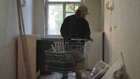 Photo for Homeless man is standing in the room of an abandoned building in search for useful things. Shopping cart of his belongings stands by his side. Homelessness and poverty, unemployment, crisis. - Royalty Free Image