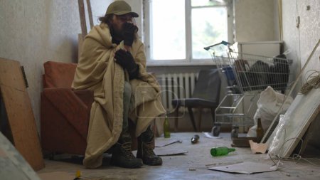 Foto de Pobre vagabundo sentado en una habitación de un edificio abandonado. Está tratando de mantener el calor debajo de una manta, bebiendo bebida caliente de una taza. Desamparo y pobreza, desempleo, crisis. - Imagen libre de derechos