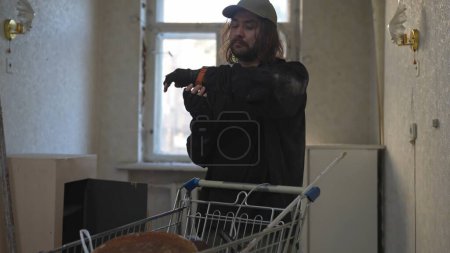 Photo for Homeless man standing is the room of an abandoned building searching for useful things in a shopping cart of his belongings, finding and wearing a watch. Homelessness and poverty, unemployment, crisis - Royalty Free Image