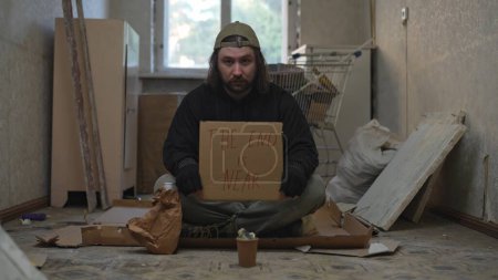 Photo for Homeless poor man sitting in a room of an abandoned building with a money jar. He is holding a piece of cardboard, the end is near is written on it. Homelessness and poverty, unemployment, crisis. - Royalty Free Image