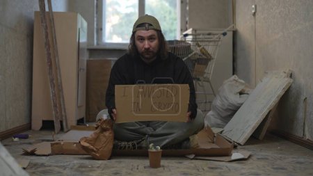Foto de Pobre vagabundo sentado en una habitación de un edificio abandonado con un frasco de dinero. Él está sosteniendo un pedazo de cartón, invertí en cripto está escrito en él. Desamparo y pobreza, desempleo - Imagen libre de derechos