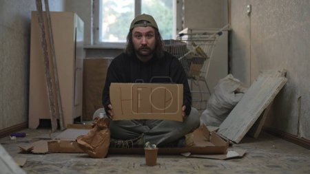 Téléchargez les photos : Pauvre sans-abri assis dans une pièce d'un immeuble abandonné avec un bocal d'argent. Il demande de l'aide avec une pancarte en carton dans les mains. Sans-abri et pauvreté, chômage, crise. - en image libre de droit