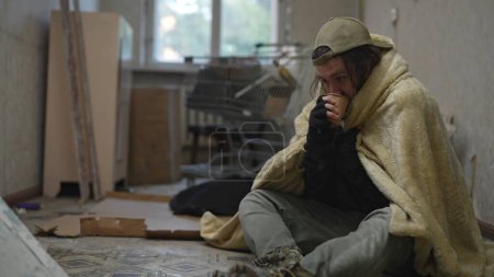 Téléchargez les photos : Pauvre sans-abri assis dans une pièce d'un immeuble abandonné. Il essaie de garder au chaud sous une couverture, en buvant une boisson chaude dans une tasse. Sans-abri et pauvreté, chômage, crise. - en image libre de droit