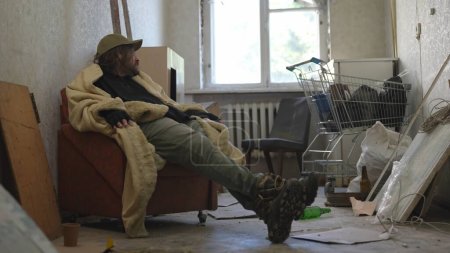 Foto de Desamparado pobre hombre cubierto de manta sentado en una habitación de un edificio abandonado lleno de sus escasas pertenencias. Está mirando por la ventana. Desamparo y pobreza, desempleo, crisis. - Imagen libre de derechos