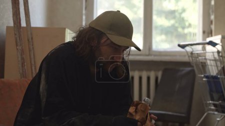 Photo for Homeless poor man sitting in a room of an abandoned building filled with his meager belongings. He is holding a bottle of alcohol in his hand. Homelessness and poverty, unemployment, crisis. - Royalty Free Image
