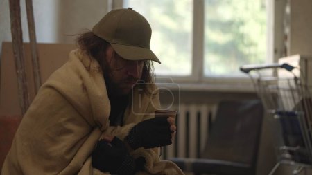 Photo for Homeless poor man sitting in a room of an abandoned building. He is trying to keep warm under a blanket, holding a cup with warm beverage. Medium shot. Homelessness and poverty, unemployment, crisis. - Royalty Free Image