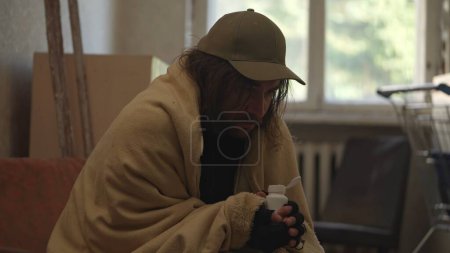 Photo for Homeless poor man covered in a blanket sitting in a room of an abandoned building filled with his meager belongings. He is holding a bottle of pills. Homelessness and poverty, unemployment, crisis. - Royalty Free Image