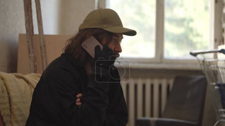 Photo for Homeless poor man sitting in a room of an abandoned building filled with his meager belongings. He is talking on the phone with somebody. Medium shot. Homelessness and poverty, unemployment, crisis. - Royalty Free Image