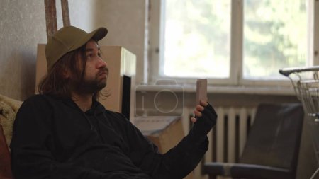 Photo for Homeless poor man sitting in a room of an abandoned building filled with his meager belongings. He is holding a phone as if he is having a videocall. Medium. Homelessness and poverty, unemployment. - Royalty Free Image