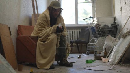 Photo for Homeless poor man covered in a blanket sitting in a room of an abandoned building filled with his meager belongings. He is holding a bottle of pills. Homelessness and poverty, unemployment, crisis. - Royalty Free Image