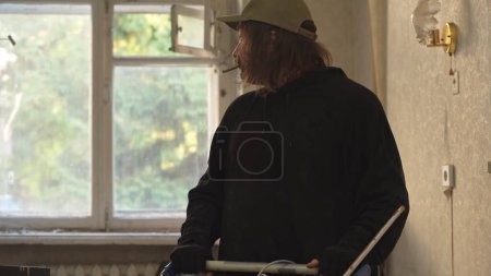 Photo for Homeless poor man standing in a room of an abandoned building, smoking a cigarette or a joint, blunt and looking at the window behind him. Medium size. Homelessness and poverty, unemployment. - Royalty Free Image