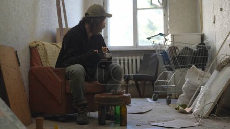 Téléchargez les photos : Pauvre sans-abri assis dans une pièce d'un immeuble abandonné. Il mange de la nourriture par la vaisselle jetable, regardant par la fenêtre. Sans-abri et pauvreté, chômage, crise. - en image libre de droit