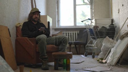 Téléchargez les photos : Pauvre sans-abri assis dans une pièce d'un immeuble abandonné remplie de ses maigres biens. Il tient un smartphone en main, surfant en ligne. Sans-abri et pauvreté, chômage, crise. - en image libre de droit
