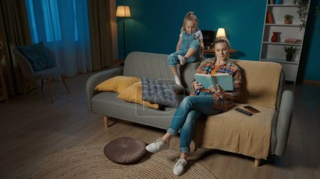 Photo for Little girl is bored near mom who is reading a book and not paying attention to her. Mom and daughter sitting on the couch in the living room - Royalty Free Image