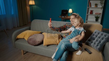 Téléchargez les photos : Mère et fille heureuses prenant selfies sur smartphone tout en étant assis sur le canapé dans le salon à la maison. Petite fille mignonne s'assoit dans les bras des mamans et sourit tout en posant - en image libre de droit