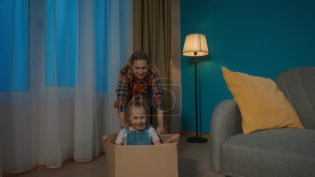 Photo for Mom and little daughter having fun. Mom is pushing a cardboard box on the floor, in which the little girl is sitting - Royalty Free Image