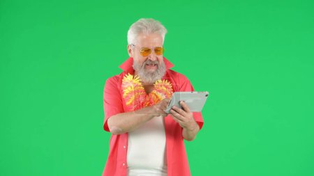 Téléchargez les photos : Concept créatif moderne pour seniors. Portrait de hipster homme âgé sur fond Chroma écran vert clé, homme tenant tablette regarder la vidéo souriant. Espace publicitaire, maquette de l'espace de travail. - en image libre de droit