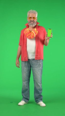 Photo for Creative modern seniors concept. Portrait of senior hipster on Chroma key green screen, man holding smartphone smiling looking at the camera. Advertising area, workspace mockup. Vertical photo. - Royalty Free Image