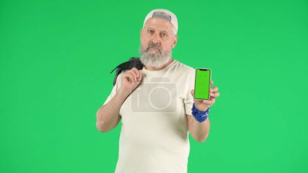 Photo for Creative modern seniors concept. Portrait of senior man hipster on Chroma key green screen background, man in white t-shirt holding smartphone looks cool at camera. Advertising area, workspace mockup. - Royalty Free Image