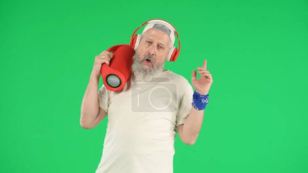 Photo for Creative modern seniors concept. Portrait of senior man hipster on Chroma key green screen background, man in white t-shirt and headphones holding music speaker. Advertising area, workspace mockup. - Royalty Free Image