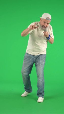 Foto de Concepto de personas mayores creativas. Retrato de hombre mayor hipster en la pantalla verde clave de Chroma, hombre en camiseta blanca cantando en el micrófono de karaoke en la cámara.. Área de publicidad, maqueta de espacio de trabajo. Foto vertical. - Imagen libre de derechos
