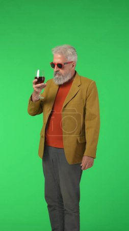 Téléchargez les photos : Concept créatif moderne pour seniors. Portrait de hipster élégant senior sur fond d'écran vert clé Chroma, homme élégant tenant un verre de vin. Espace publicitaire, maquette d'espace de travail. Photo verticale. - en image libre de droit