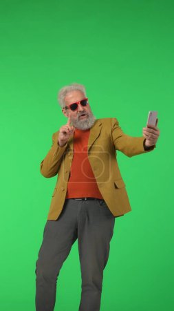 Téléchargez les photos : Concept créatif moderne pour seniors. Portrait hipster élégant senior sur fond d'écran vert clé Chroma, homme élégant posant pour selfie sur smartphone. Espace publicitaire, maquette d'espace de travail. Photo verticale. - en image libre de droit