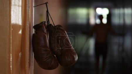 Photo for In the close up shot, brown leather boxing gloves hang on a cord from a rusty nail on a door frame. In the background, a man is walking down the corridor. He goes for the gloves, he is a boxer - Royalty Free Image