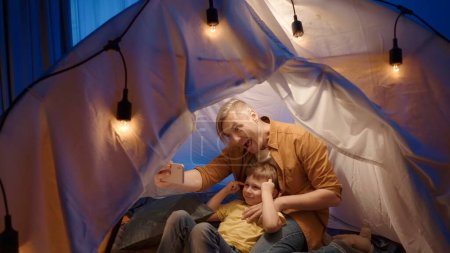 Téléchargez les photos : Un père et son fils sont assis sur des peaux de fourrure dans une tente dans un salon sombre et utilisent un smartphone. Homme et garçon prennent des selfies ensemble, faisant des grimaces drôles, souriant, gros plan - en image libre de droit