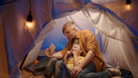 Photo for A father and son sit on fur skins in a tent in a dark living room and use a smartphone. Man and boy take selfies together, making funny faces, smiling, close up - Royalty Free Image